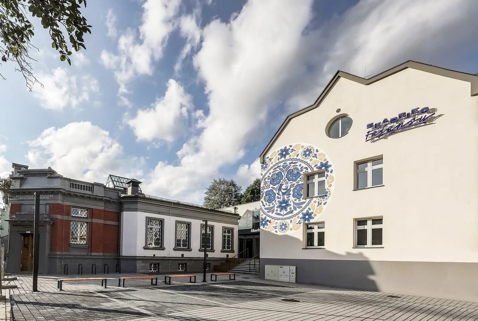 A modern white building with a large blue and yellow circular floral mural and a sign in blue, adjacent to an older brick and white building, both under a cloudy blue sky.